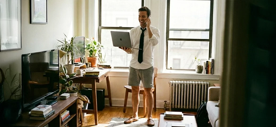 Uzaktan Çalışanlar Nasıl Terfi Alır?: Ev ortamında üstü gömlek altı şortla çalışan mutlu bir ofis çalışanı ve Uzaktan Çalışanlar Nasıl Terfi Alabilir yazı başlığı.